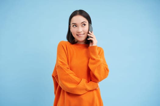 Korean girl in orange sweater talks on mobile phone, speaks on cellphone with happy smiling face, orders delivery, consults with someone on telephone.