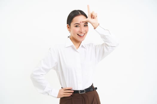 Successful businesswoman looks confident, shows L letter, loser sign on forehaed, feels proud of herself, smiles pleased over white background.