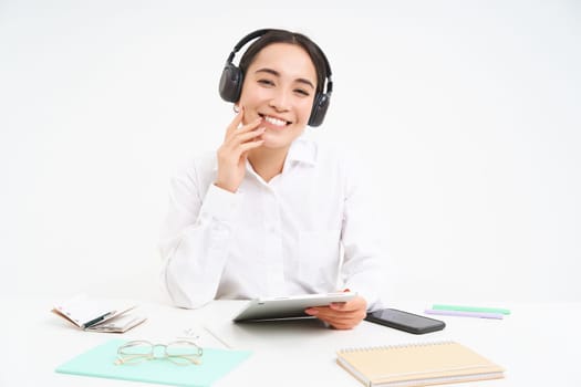 People and workplace. Smiling asian woman sits in office, student listening music in headphones and holding digital tablet, white background.