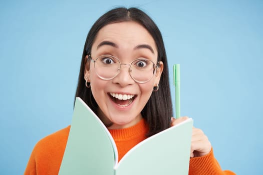 Amazed young asian woman in glasses, has an idea, writes down with pen in notebook, has revelation, enjoys listening course, student studying, blue background.