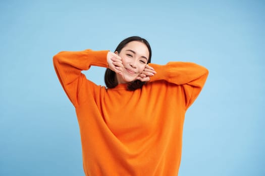 Smiling young korean woman stretching her arms with satisfied face, pleased after good nap, waking up, standing over blue background.