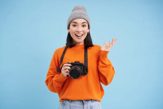 Stylish asian girl with digital camera, taking pictures. Woman photographer smiling, standing over blue background.