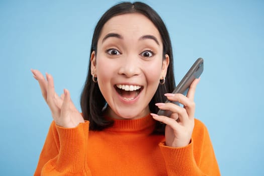 Girls reaction to amazing news she received on mobile phone. Happy asian woman looks surprised and excited, blue background.