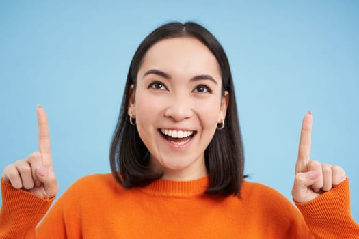 Beautiful asian girl points fingers up, shows banner on top, advertisement, stands over blue background. Copy space