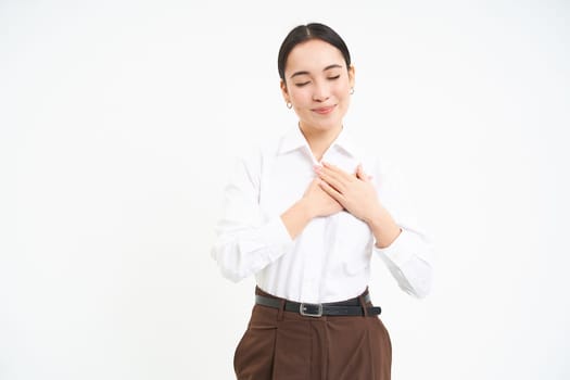 Romantic young asian woman holds hands on her heart with dreamy smile and eyes closed, white background.