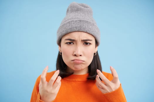 Close up portrait of annoyed asian girl, shakes hands and sulks, looks angry and irritated, stands over blue background. Negative emotions