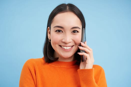 Technology concept. Close up of smiling asian woman talks on mobile phone, having conversation on cellphone, blue background.