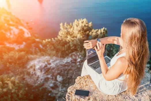 Successful business woman in yellow hat working on laptop by the sea. Pretty lady typing on computer at summer day outdoors. Freelance, travel and holidays concept.