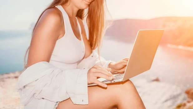 Digital nomad, Business woman working on laptop by the sea. Pretty lady typing on computer by the sea at sunset, makes a business transaction online from a distance. Freelance remote work on vacation