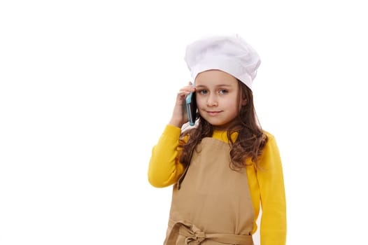 Beautiful child girl, little baker confectioner wearing white chef's hat and beige apron, talking on mobile phone, smiling at camera. Food delivery service. Advertising shot on white studio background