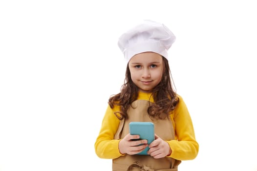 Lovely baby girl dressed as chef confectioner, smiles looking at camera, holds a modern smartphone over white isolated studio background. Mobile apps. Food ordering. Culinary. Children and technology