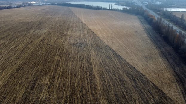 Large plowed field with plowed land and yellow dry straw after harvesting wheat, a lake, a highway with driving cars on an autumn day. Agro industrial agricultural farm landscape. Aerial drone view.