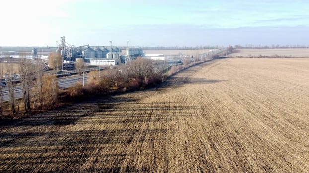 Agricultural industrial landscape. Plowed field after harvesting dry wheat, an elevator, a highway with moving cars on an autumn sunny day. Yellow field with dug up earth, road, cars, grain elevator