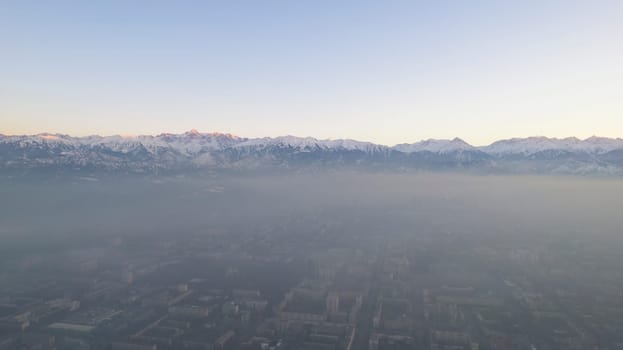 Epic gray smog is visible at sunset over the city. A bird's-eye view from a drone of houses, roads, cars and parks. White clouds and snowy mountains are illuminated by orange rays of the sun. Almaty