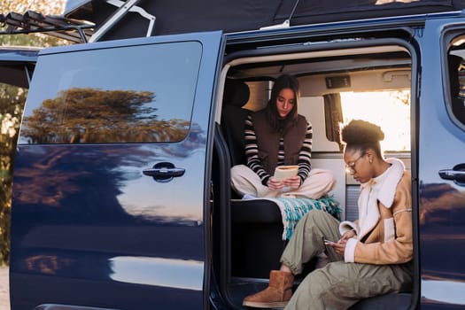 two young women enjoying camper van life reading at sunset, concept of travel adventure and female friendship