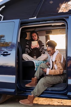 two young women enjoying camper van life reading at sunset, concept of travel adventure and female friendship