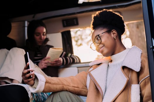 smiling african woman taking selfie photo with mobile phone in camper van, concept of adventure travel with friend and technology of communication