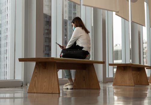 Bangkok,Thailand - Aug 13, 2022 - A calm young asia woman sitting on Modern wood bench wearing face mask protection for virus coronavirus covid 19 and using smart phone browse internet reading media news in empty hall room with glossy floor and cityscape. Side view, No focus, specifically.