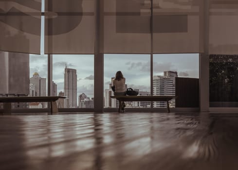 Bangkok,Thailand - Aug 13, 2022 : A calm young asia woman sitting on Modern wood bench looking through transparent wall in empty hall room with glossy floor and cityscape with skyscrapers. Rear view, Selective focus.