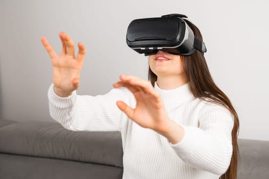 Curious woman swipes virtual interface with hands looking through VR glasses. Young housewife sits on stylish sofa enjoying visual effects at home