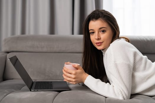 Portrait of a beautiful young girl lying on a sofa with a cup of tea and a laptop.
