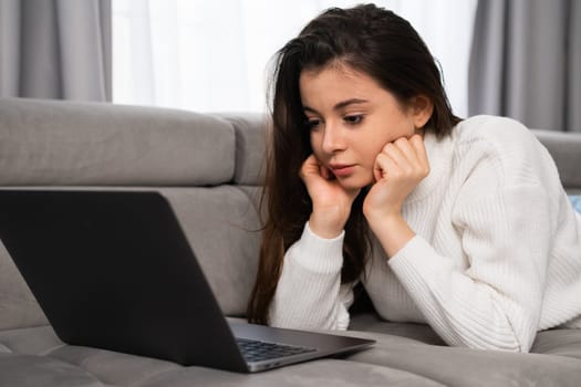 Satisfied woman relaxing on comfortable couch using laptop at home and watching video on laptop