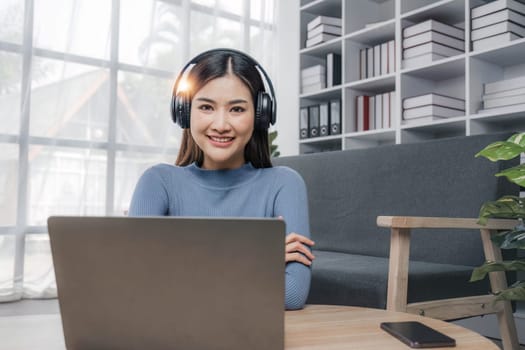 Smiling young asian woman using laptop web camera while wearing headphones and sitting on the rug beside to the sofa at homes..