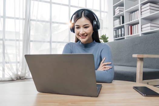 Smiling young asian woman using laptop web camera while wearing headphones and sitting on the rug beside to the sofa at homes..