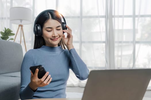 Smiling young asian woman using laptop web camera while wearing headphones and sitting on the rug beside to the sofa at homes..