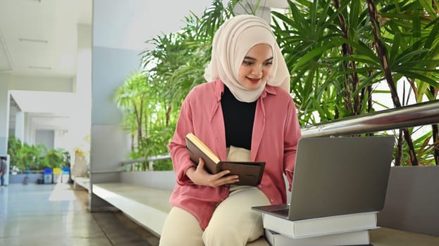 Smiling Asian muslim student holding book and searching information on laptop. Education and Technology concept.