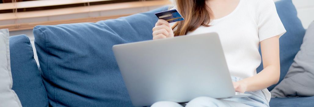 Young asian woman sit on sofa using laptop computer shopping online with credit card buying to internet, happy girl payment with e-business on couch, purchase and payment, business concept.