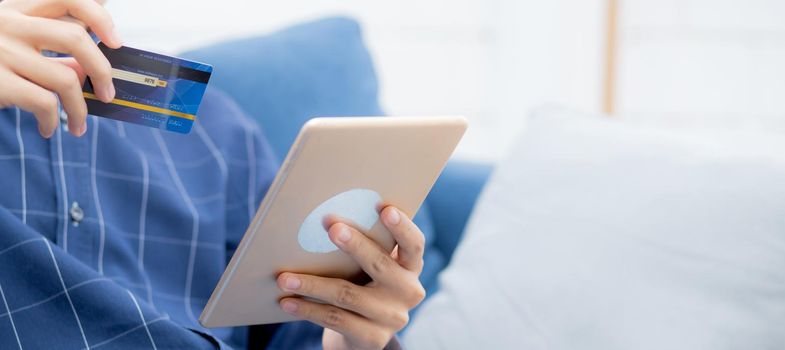 Young asian man smiling holding credit card shopping online with tablet computer buying and payment, male using debit card purchase or transaction of finance, lifestyle and e-commerce concept.