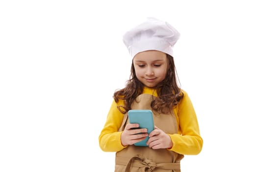 Beautiful child girl, little baker confectioner wearing white chef's hat and beige apron, checking smartphone, enyoying new mobile application. Food delivery service. Advertising shot white background