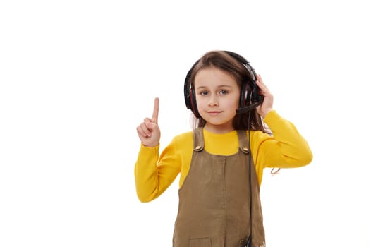 Beautiful little girl, first grader wearing headphones for online education or distance learning, pointing her index finger on a copy space, smiling looking at camera, isolated on white background.