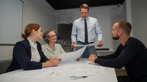 A caucasian man stands and holds a drawing, three colleagues sit at a table and listen to him