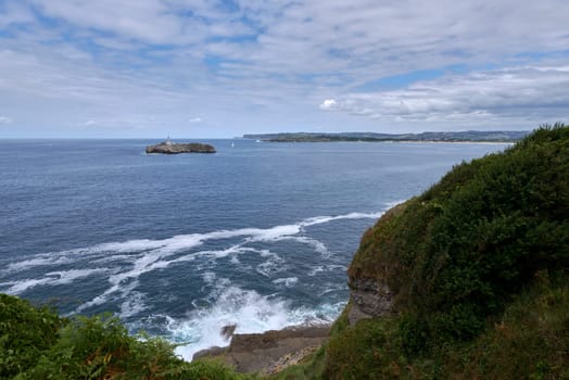 A lighthouse on the Cantabrian Sea coast, and coast line, Calm waters, waves on the shore, vegetation, cliffs, sailboats, seagulls