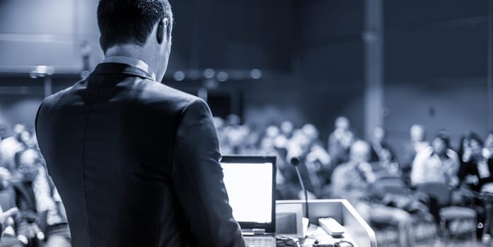 Speaker giving a talk on corporate business conference. Unrecognizable people in audience at conference hall. Business and Entrepreneurship event. Blue toned grayscale image.