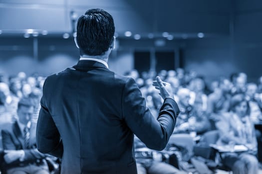 Speaker giving a talk on corporate business conference. Unrecognizable people in audience at conference hall. Business and Entrepreneurship event. Blue toned grayscale image.