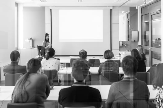 Female speaker giving presentation in lecture hall at university workshop. Rear view of unrecognized participant in audience. Scientific conference event. Black and white image.
