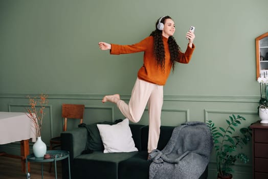 A happy, pretty young woman wearing headphones dances to her favorite song at home. Cheerful funny girl listening to popular music, moving, jumping on the couch in the living room, experiencing joy