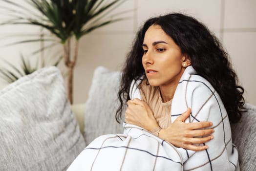 A beautiful young Arab woman escapes from the cold by wrapping herself in a blanket. The woman sat on a gray sofa and wrapped herself in a blanket with her hands holding her shoulders. Young girl trying to keep warm in her room. A beautiful Muslim woman with dark curly hair looks down in front of her in puzzlement. She is concerned about the temperature in her apartment. The cold affects her health and she starts to get sick.