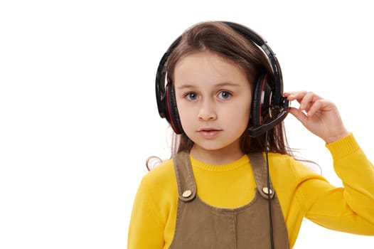 Adorable 5-6 years Caucasian child, lovely little girl wearing headphones for online education or distance learning, looking at camera, isolated on white background. Close-up portrait of smart pupil