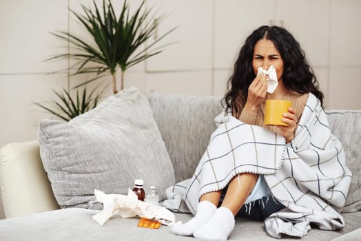 A young girl caught a cold and is being treated at home. Woman blowing her nose and wiping her nose with a tissue.Sitting on a gray sofa covered with a blanket and drinking a hot drink.