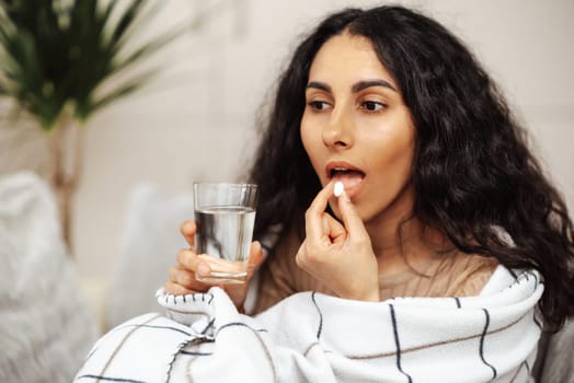 A beautiful young girl of Arab appearance takes a pill in one hand, she holds a pill in the other glass of water. The woman is in a homely cozy atmosphere and wrapped in a blanket. Taking a pill, a woman takes care of her health. Any virus, allergy or disease is not scary when there is the right treatment.