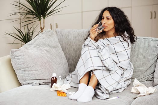 Woman is sitting on a gray sofa wrapped in a blanket around her with a lot of medicines and handkerchiefs. In one hand she holds a glass and drink water. On the face the emotion of anxiety puzzled.
