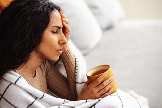 A young beautiful Arab woman is sitting on a gray sofa wrapped in a blanket and drinking tea. Stress and problems caused a severe headache. From a severe migraine, the woman closed her eyes.