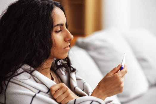 A young beautiful Arab woman holds a thermometer with one hand and holds a plaid with the other while sitting on a gray sofa. A woman looks puzzled at a thermometer in a homely atmosphere.