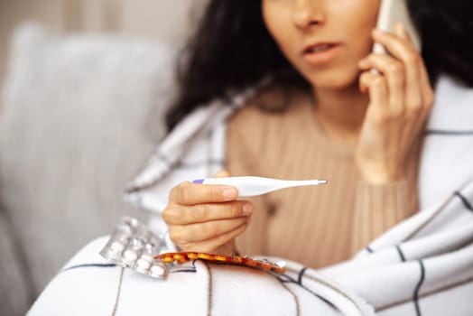 Young woman calling family doctor and consulting about treatment. A woman in one hand holds a smartphone in the other electronic thermometer. In the foreground, the thermometer in the background, the woman herself, along with the phone, is out of focus and fragmentary. A woman sits in her room on a gray sofa wrapped in a blanket. She feels that she is ill and the call to the doctor is a consultation about her treatment.