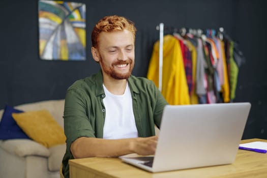 Caucasian man with red hair, sitting at desk in cozy home interior. He work on laptop, emphasizing concept of freelance work and ability to work from home. Man using his laptop to access his work remotely. High quality photo