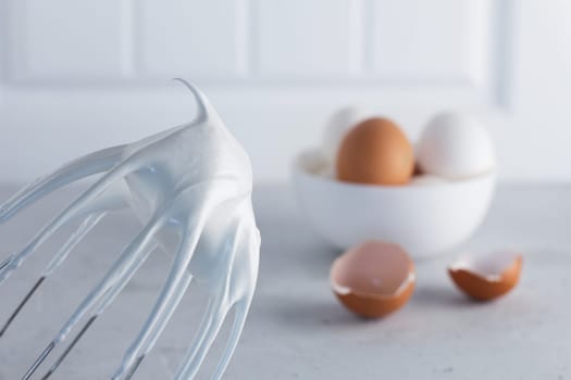 Whisked egg whites - whipped Italian meringue on a wire whisk and eggs on a gray background. copy space.